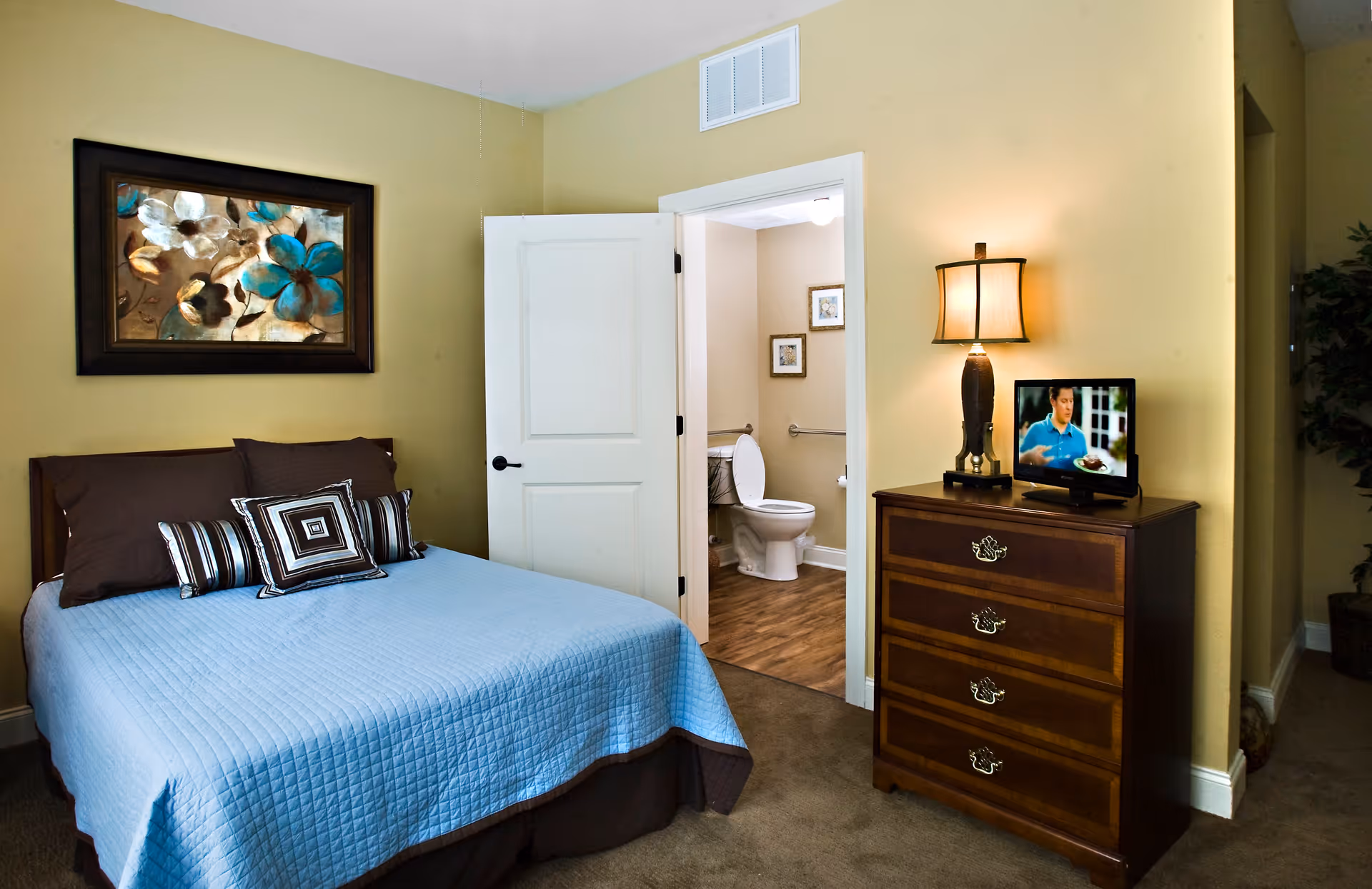 A bedroom with a bed covered in a blue quilt and multiple pillows. There is a framed floral painting on the wall above the bed. An open door reveals a bathroom with a toilet and framed pictures on the wall. A wooden dresser with a lamp and a small television is positioned near the door.