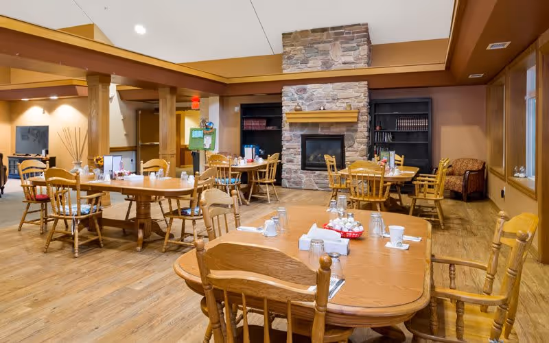 A cozy dining room with wooden tables and chairs arranged neatly. The room features a stone fireplace in the center background with built-in shelves on either side. The tables are set with glasses, napkins, and condiments. The floor is wooden, and the walls are painted in warm tones, creating a welcoming atmosphere.