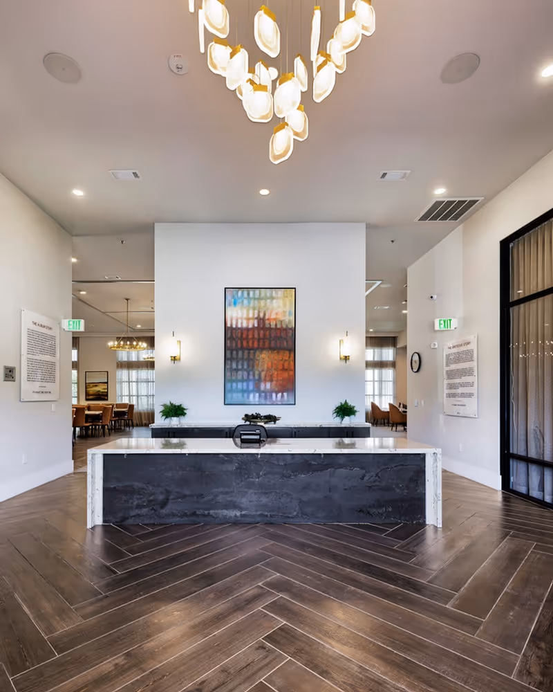 Modern interior space with a large marble reception desk in the center, dark wood herringbone flooring, a colorful abstract painting on the white wall behind the desk, two wall sconces, and a contemporary chandelier hanging from the ceiling. There are exit signs and seating areas visible in the background.