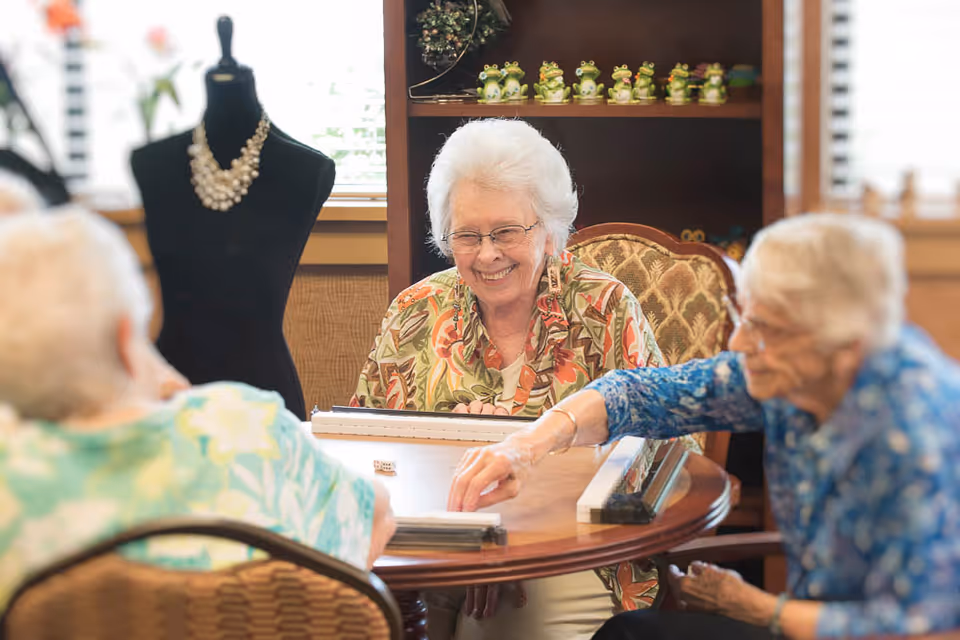 Three elderly women sitting around a wooden table playing a game with dice. One woman in the center is smiling and wearing glasses and a floral patterned shirt. The background shows a shelf with decorative frog figurines and a black mannequin with a necklace.