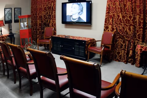Small screening/activity room with rows of chairs facing a wall-mounted TV, red patterned curtains, and a popcorn machine.