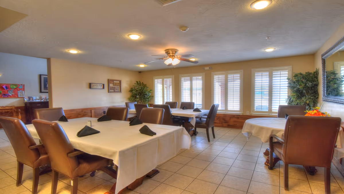 A dining room with several tables covered in white tablecloths and set with black folded napkins. The room has brown leather chairs, tiled floor, large windows with white shutters, a ceiling fan with lights, and decorative plants in the corners.
