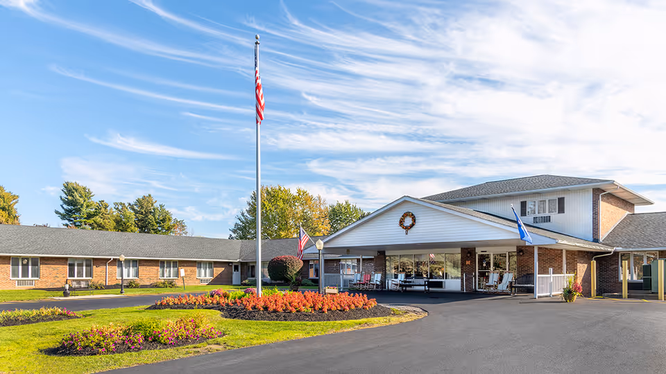 Exterior view of Elderwood Village at Bassett Park, showing a single-story brick building with a covered entrance, an American flag on a tall flagpole, well-maintained flower beds, and a clear blue sky with wispy clouds.