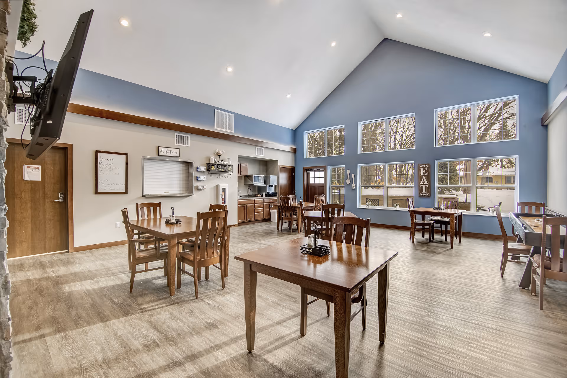 A bright dining room with several wooden tables and chairs arranged neatly. The room features large windows letting in natural light, blue accent walls, and a high vaulted ceiling with recessed lighting. There is a small kitchen area with cabinets, a microwave, and a serving window. A wall-mounted TV is visible on the left side.