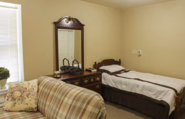 Small bedroom with a twin bed, wooden dresser and mirror, and a plaid loveseat by a window.