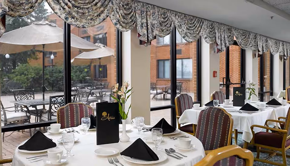 A bright dining room with round tables set with white linens and black napkins beside large windows overlooking an outdoor patio.
