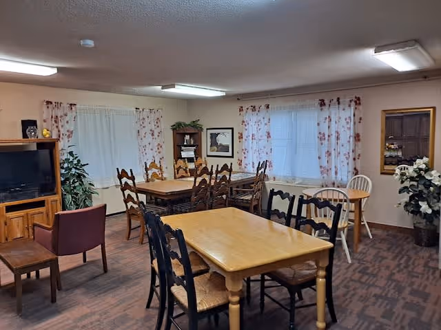 A cozy common area with multiple wooden tables and chairs arranged for seating. The room has two windows with floral curtains, a television on a wooden stand, a small bookshelf, and some potted plants. The floor is carpeted and the ceiling has fluorescent lighting.