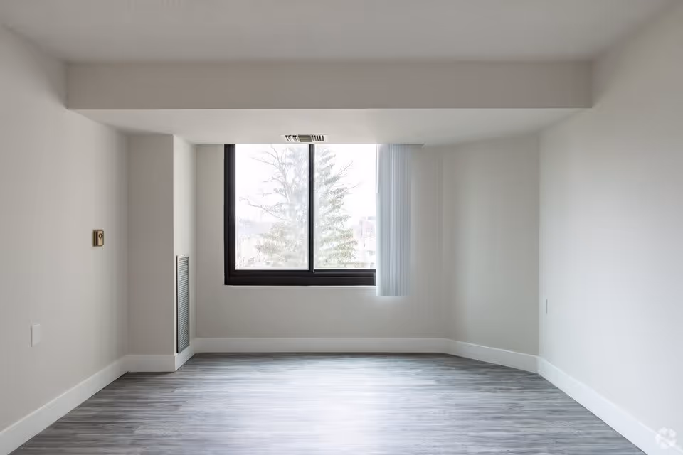 Empty room with light gray walls, a large window with vertical blinds partially open, and gray wood-style flooring. The window shows a view of a tree outside.