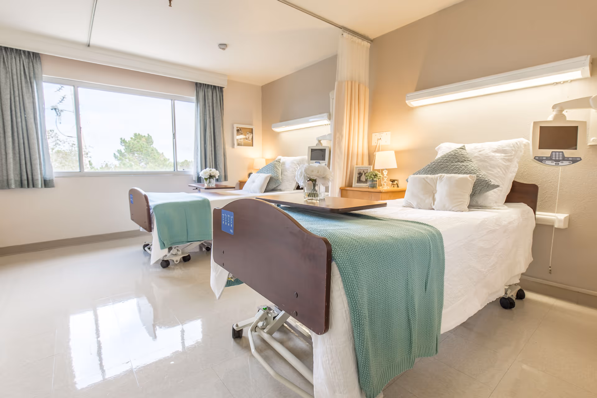 A bright and clean room with two hospital-style beds, each made with white linens and light green blankets. The beds have adjustable headboards and are equipped with medical monitors. There is a large window with curtains letting in natural light, and a small wooden nightstand with a lamp and decorative items between the beds. The floor is shiny and reflective, and the walls are painted in neutral tones.