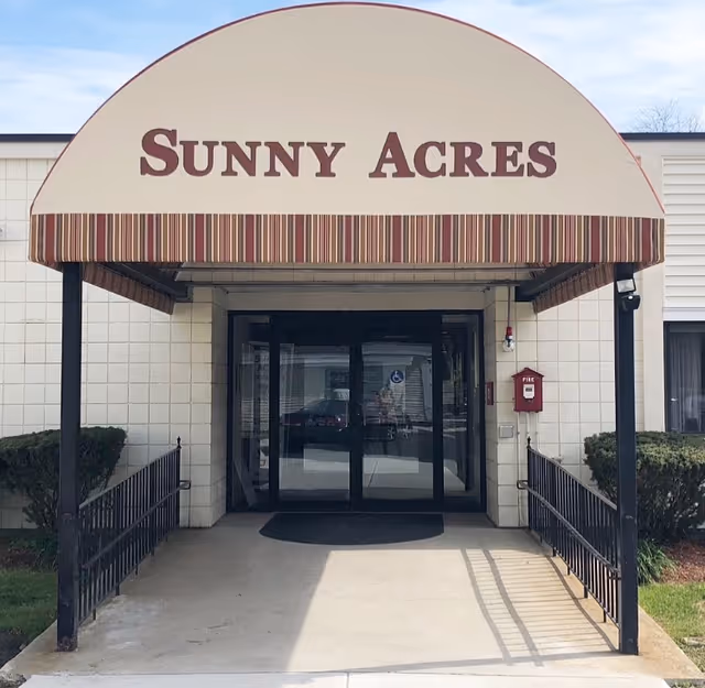 Entrance to Sunny Acres facility with a beige and striped awning displaying the name Sunny Acres above double glass doors. There are black metal railings on both sides of the concrete walkway leading to the entrance, with bushes on either side.