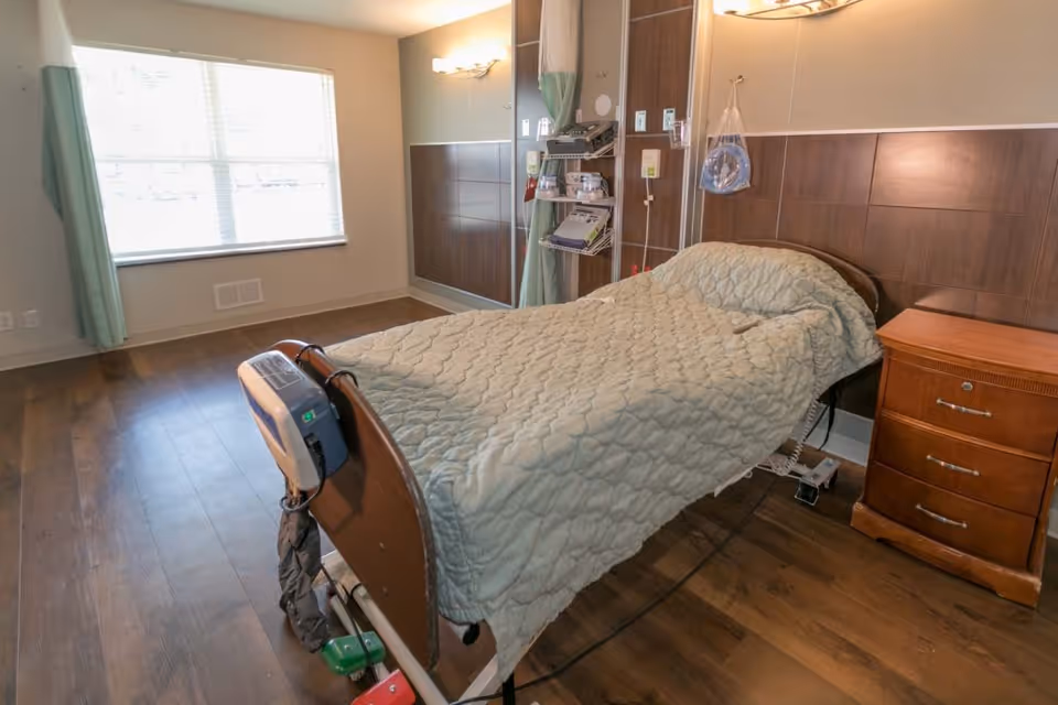 A single hospital-style bed with a quilted beige blanket in a room with wooden flooring and a large window with green curtains. There is medical equipment attached to the bed and a wooden nightstand with drawers beside it. The walls have wood paneling halfway up and a light fixture above the bed.