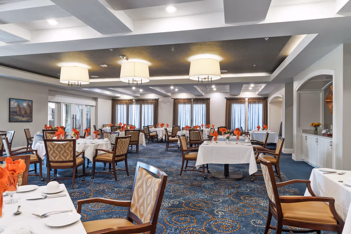 A spacious dining room with multiple round tables covered with white tablecloths and set with plates, silverware, and orange folded napkins. The room features patterned carpet with blue and orange circular designs, upholstered chairs, large windows with sheer curtains, and modern ceiling lights.