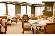 A bright dining room with multiple tables covered in white tablecloths, each set with placemats and small floral centerpieces. The room has large windows allowing natural light to fill the space, green walls, and wooden chairs with cushioned seats arranged around the tables.