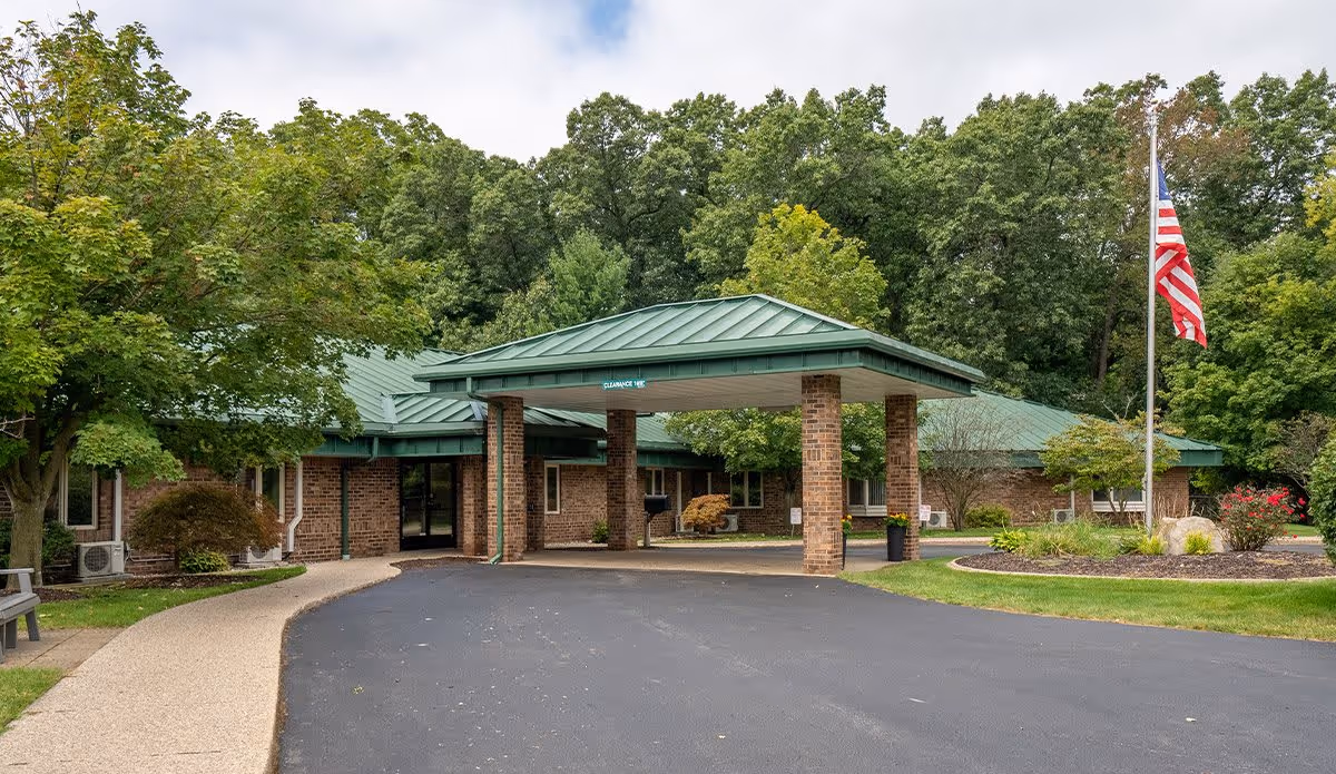 Exterior view of a single-story brick building with a green metal roof and a covered entrance supported by brick columns. There is a paved driveway leading to the entrance, surrounded by green grass, trees, and landscaping. An American flag is flying on a flagpole to the right of the entrance.
