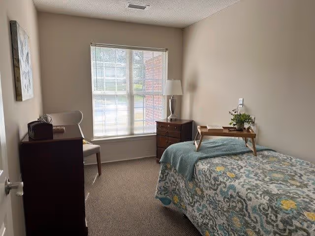 A small, neatly arranged bedroom with a single bed covered in a patterned quilt. A wooden tray with a book and a small plant is placed on the bed. Next to the bed is a wooden nightstand with a lamp and another small plant. A chair and a dresser are positioned near a window with blinds, allowing natural light to enter the room. The walls are painted a light beige color.