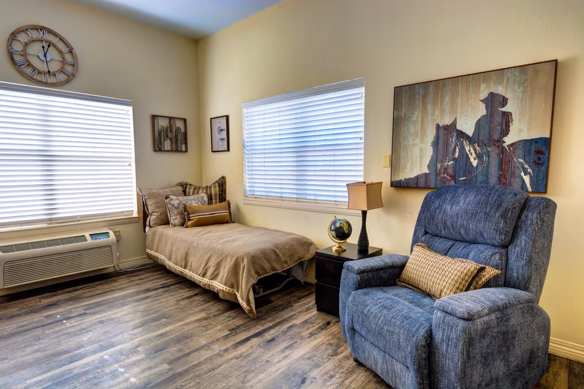 A cozy bedroom with a single bed covered in beige bedding and multiple pillows, a blue upholstered recliner chair with a patterned cushion, a small black nightstand with a globe and a lamp, two windows with white blinds, and wall art including a large painting of a cowboy on a horse and smaller framed pictures.