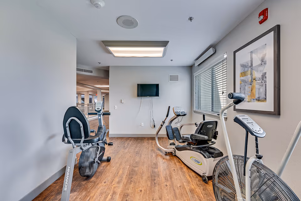 Fitness room with recumbent bikes and cardio equipment, a wall-mounted TV, window blinds, and framed artwork.