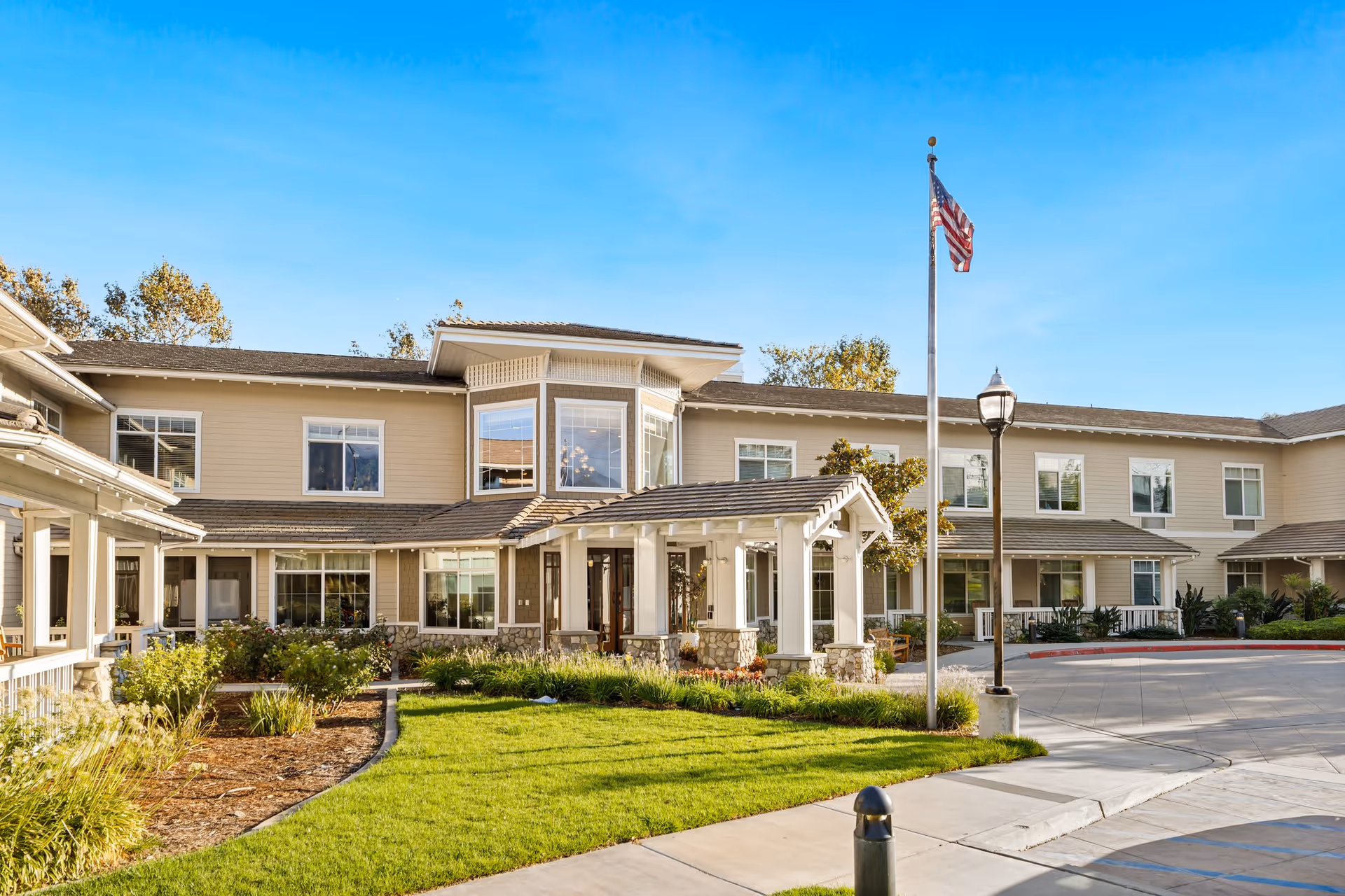 Exterior view of a two-story senior living facility building with beige siding and white trim. The entrance features a covered porch with white pillars and stone bases. There is a well-maintained lawn and landscaped garden beds in front, along with a flagpole flying the American flag and a streetlamp near the driveway.
