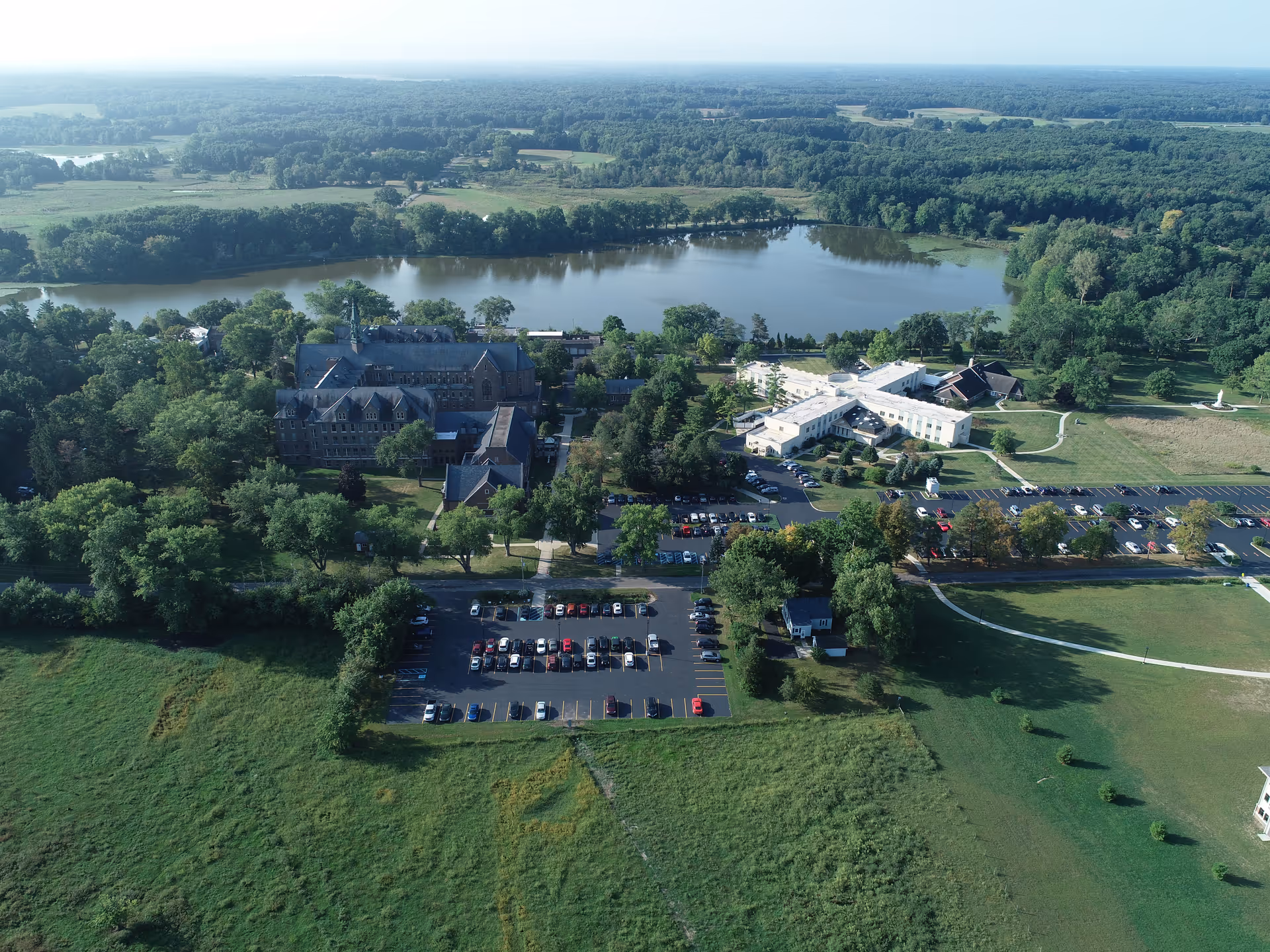 Aerial view of the Catherine Kasper Life Center facility surrounded by trees and greenery, with a large parking lot in front and a river in the background under a clear sky.