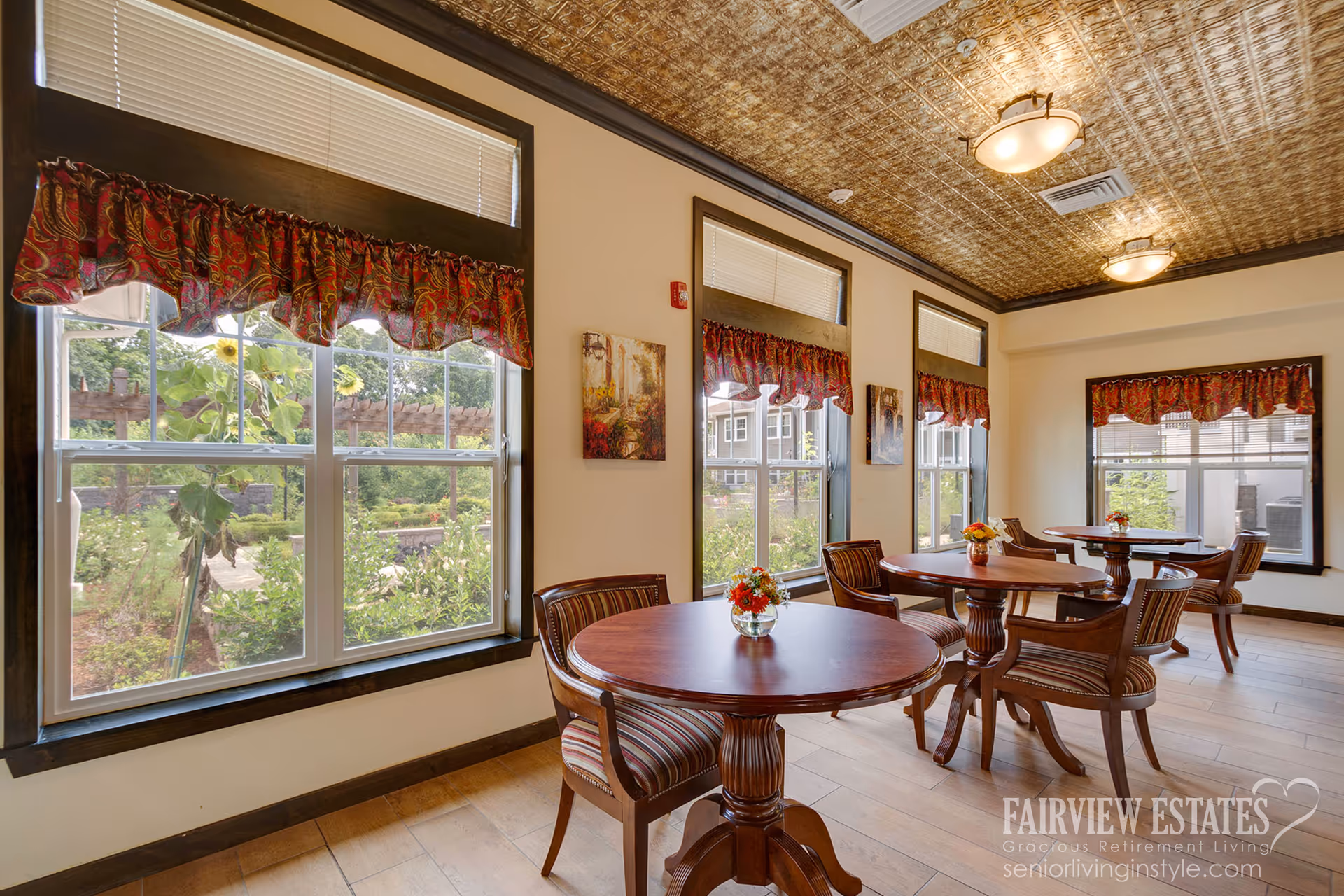 Sunlit dining area with round wooden tables and chairs by large windows overlooking a garden.