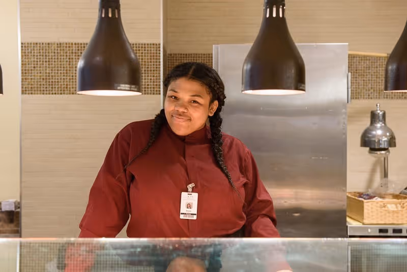 A woman with braided hair wearing a maroon uniform and an ID badge stands behind a glass counter in a kitchen or food service area with hanging black pendant lights above her.