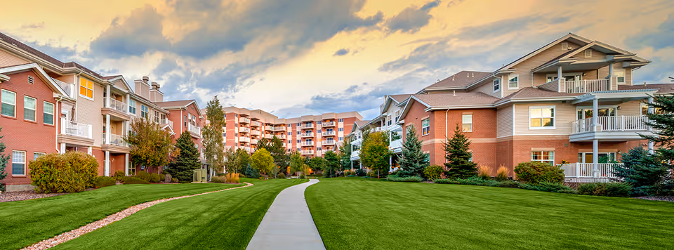 A paved walkway through a green lawn bordered by multi-story senior living buildings under a dramatic sky.
