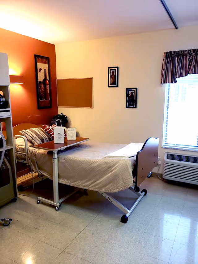 A single hospital-style bed with beige bedding and striped pillows in a room with light yellow and orange walls. There is a wooden overbed table with items on it, a corkboard on the wall, framed artwork, a window with blinds and a striped valance, and an air conditioning unit below the window.