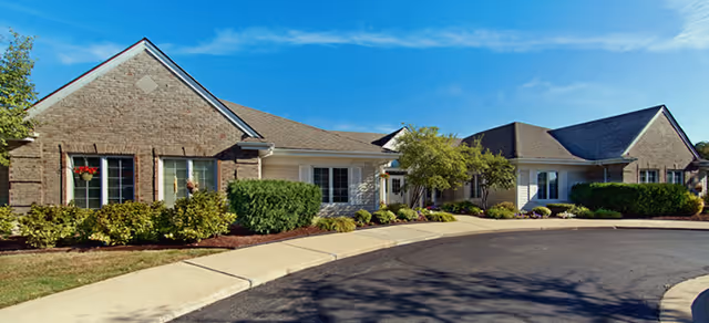 Single-story brick and siding senior living building with landscaped shrubs, trees, and a curved driveway under a blue sky.