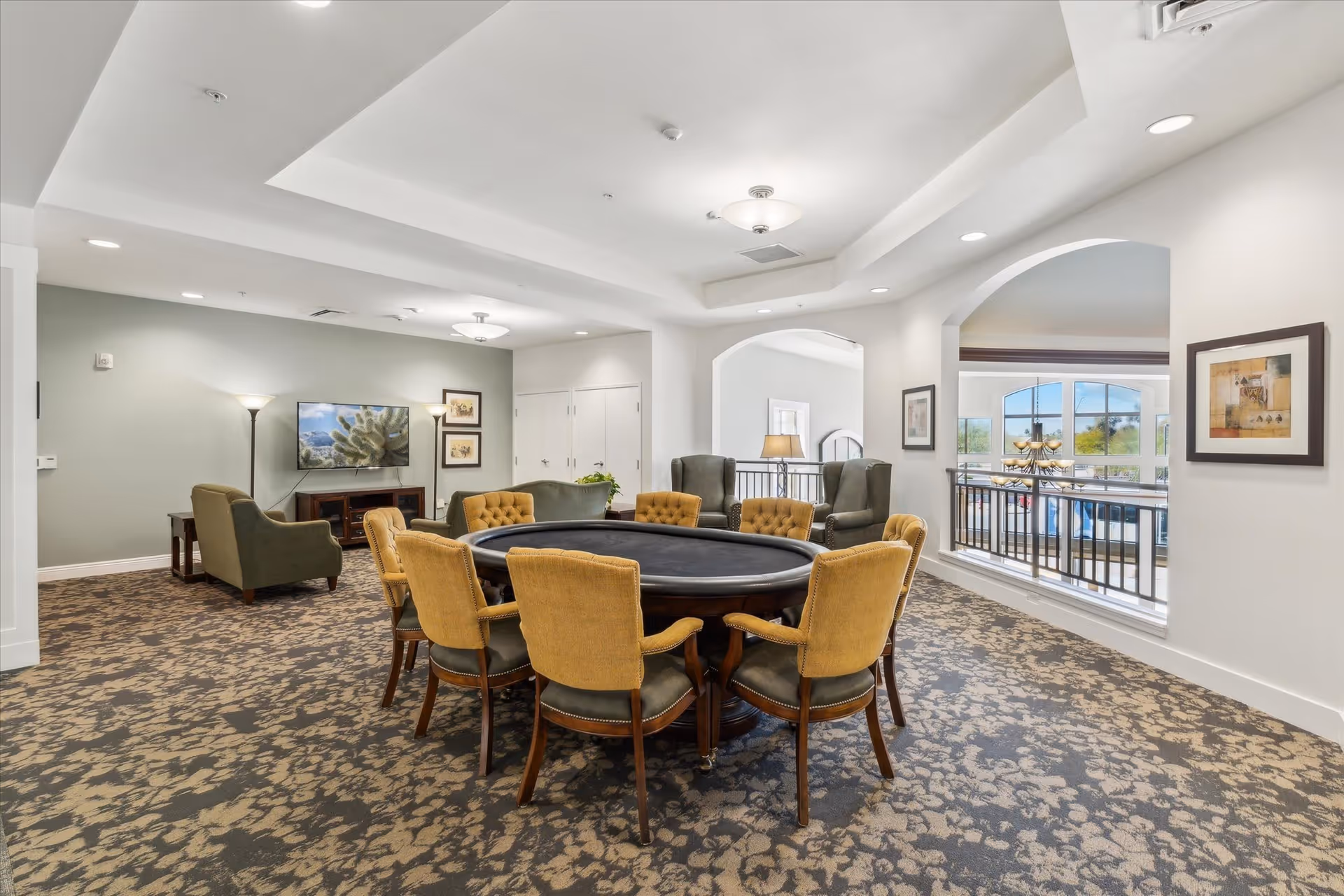 A spacious and well-lit common area in an assisted living facility featuring a large round table with eight yellow cushioned chairs around it. In the background, there are two green armchairs facing a wall-mounted TV with two floor lamps on either side. The room has patterned carpet flooring, white walls, and large arched windows overlooking an adjacent area with a chandelier.
