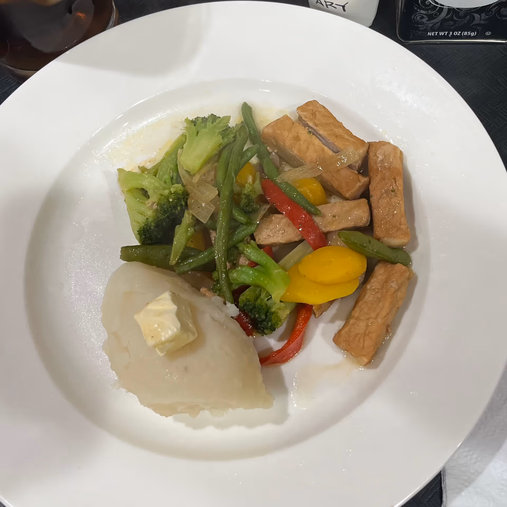 White plate with mashed potatoes topped with a pat of butter and a serving of mixed vegetables and sliced meat.