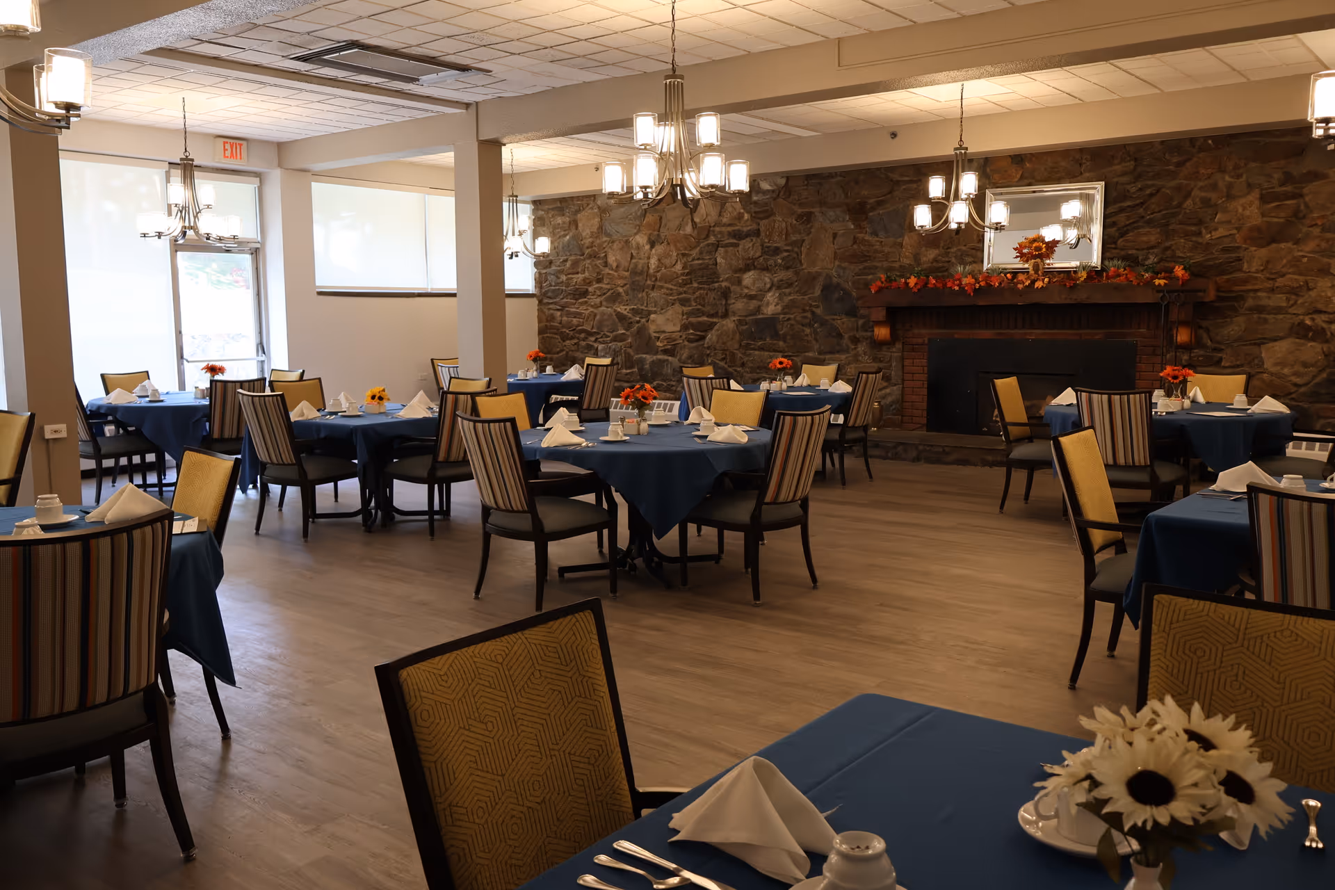 A dining room with multiple round tables covered with blue tablecloths, each set with white napkins, cups, and small flower arrangements. The room features a stone wall with a fireplace decorated with autumn-themed garlands and a mirror above it. There are chandeliers hanging from the ceiling providing warm lighting, and large windows letting in natural light.