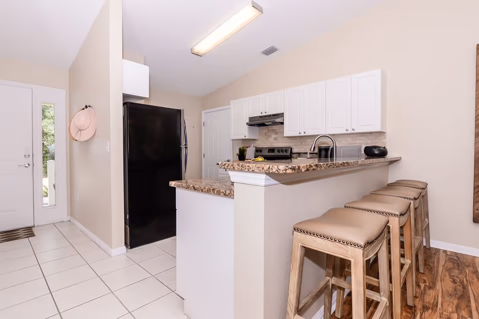 A modern kitchen with beige walls and white cabinets. There is a black refrigerator, a stove with a range hood, a microwave, and a granite countertop with a sink. Four wooden bar stools with beige cushioned seats are lined up along the counter. A pink hat hangs on the wall near a white door with a window.