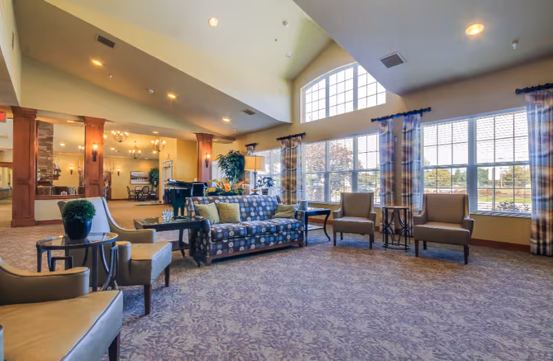 A spacious and well-lit living room area with large windows allowing natural light to fill the space. The room features a patterned sofa with green and yellow cushions, several beige armchairs, small round tables, and a carpeted floor with a floral design. The ceiling is vaulted with recessed lighting, and there are decorative wooden columns and a stone fireplace visible in the background.