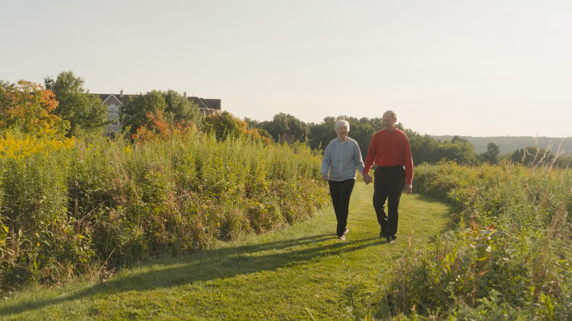 An elderly couple holding hands and walking along a grassy path surrounded by tall green plants and trees, with a building visible in the background under a clear sky.