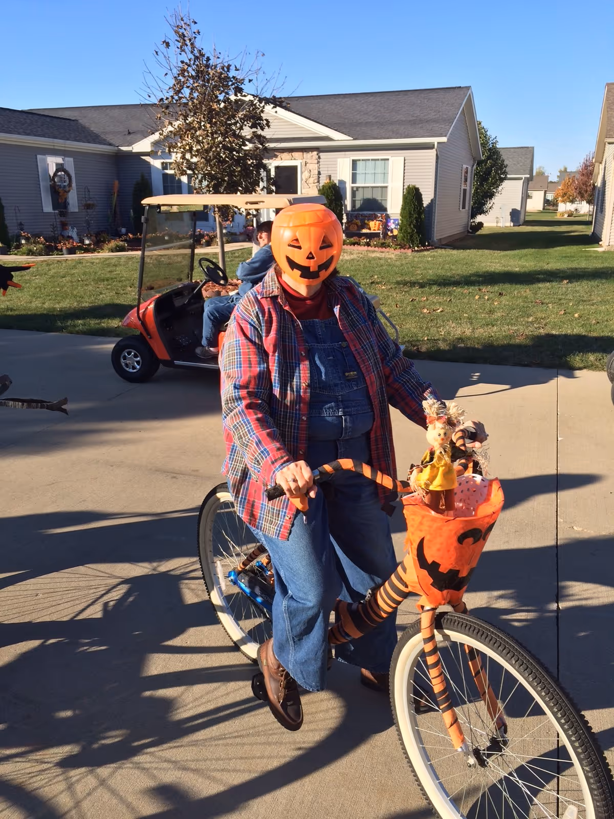 Person wearing a pumpkin mask and plaid shirt riding a bicycle decorated with Halloween-themed orange and black striped elements and a pumpkin face basket, on a driveway in front of residential houses with green lawns and a golf cart in the background.