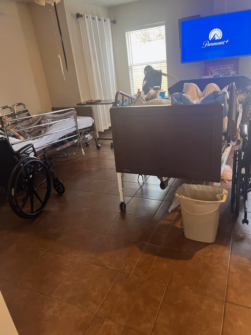 A room with two hospital beds, one occupied by a person covered with a blanket, a wheelchair, a trash bin, a window with white curtains, and a wall-mounted TV displaying the Paramount+ logo.