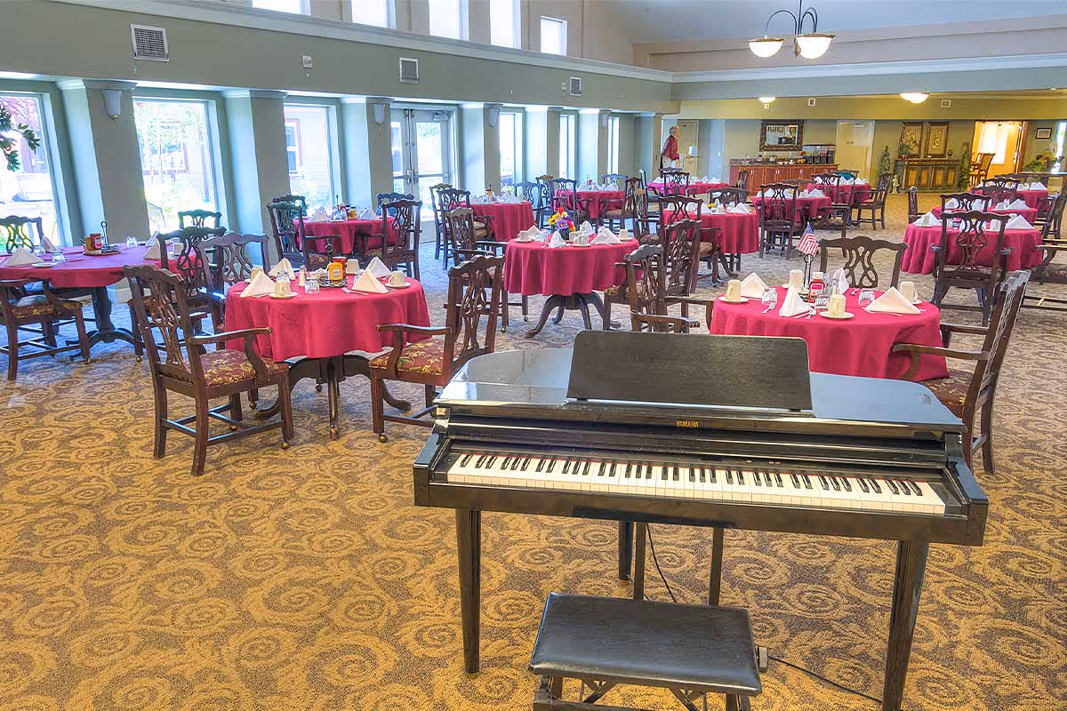Spacious dining room with round tables dressed in red tablecloths and a piano in the foreground.