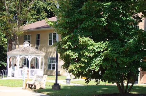 Yellow two-story building with a white gazebo, bench, and a large tree in a grassy front yard.