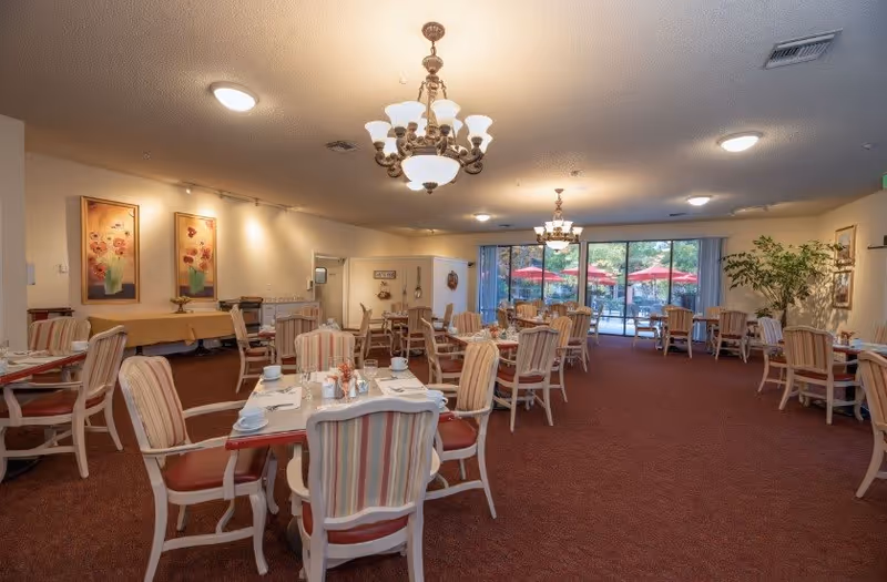 A spacious dining room with multiple tables and chairs arranged neatly. Each table is set with white cups, plates, and napkins. The room features warm lighting with chandeliers and ceiling lights. Large windows at the far end reveal an outdoor patio area with red umbrellas and greenery. The walls are decorated with framed floral paintings and there is a large potted plant on the right side.