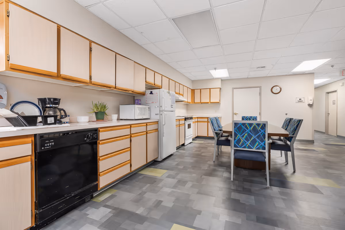 A spacious kitchen area in a senior living facility with light wood cabinets, a black dishwasher, white refrigerator, microwave, coffee maker, and a dining table with six chairs featuring blue patterned upholstery. The floor has a gray and yellow tile pattern, and the ceiling has white tiles with fluorescent lighting. A clock is mounted on the wall near a closed door.