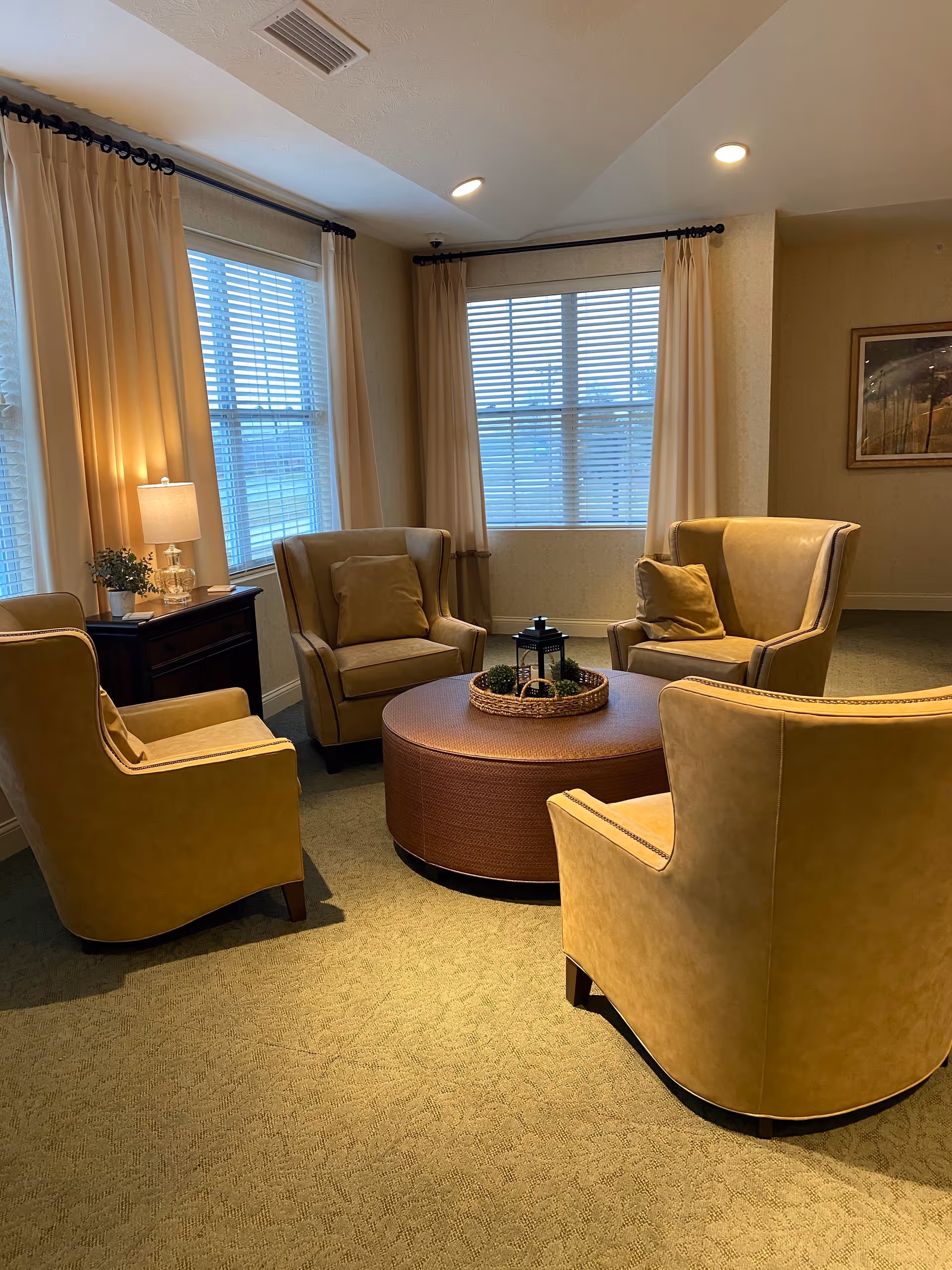A cozy seating area with four tan leather armchairs arranged around a round brown ottoman with a decorative tray and lantern on top. The room has beige walls, carpeted floor, two large windows with white blinds and cream curtains, a side table with a lamp and small plant, and a framed picture on the wall.