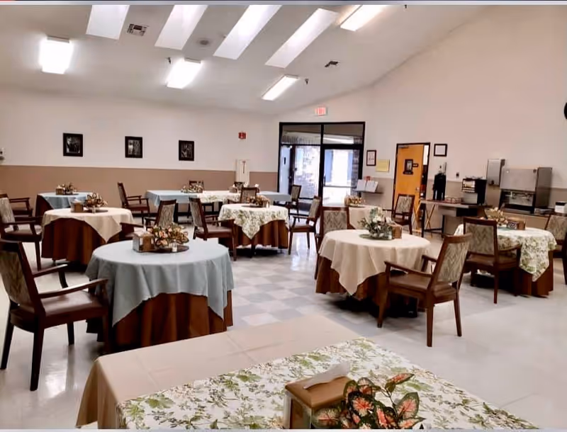 A spacious dining room with multiple round tables covered with tablecloths in various colors, each decorated with floral centerpieces. Wooden chairs with cushioned seats surround the tables. The room has a high ceiling with skylights, a tiled floor, and a door leading outside. There is a coffee and beverage station along one wall.
