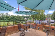 Outdoor patio area with several metal tables and chairs under large green umbrellas, surrounded by a brick wall railing. Trees and a flagpole with an American flag are visible in the background under a clear blue sky.
