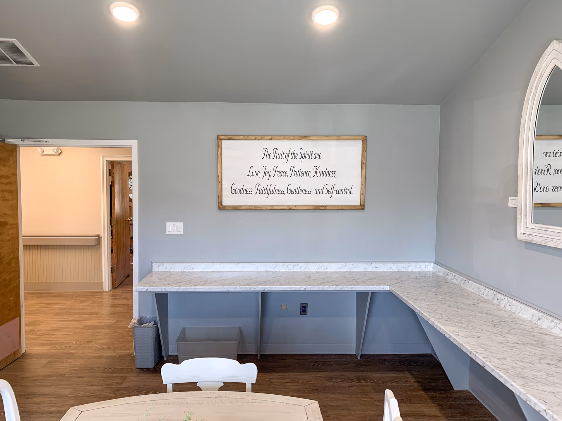 A room with light gray walls and wood flooring featuring a white marble countertop running along two walls in an L-shape. A framed sign on the wall reads 'The Fruit of the Spirit are Love, Joy, Peace, Patience, Kindness, Goodness, Faithfulness, Gentleness and Self-control.' There is a round wooden table with white chairs in the foreground, a trash bin under the countertop, and a mirror on the right wall reflecting part of the sign. An open door leads to a hallway with wood flooring and beige walls.