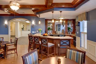 Interior view of a senior living facility common area featuring a kitchen island with bar stools, wooden ceiling beams, pendant lighting, and a seating area with striped upholstered chairs and a round table. The walls are painted blue with white wainscoting, and there is an arched doorway leading to another room.