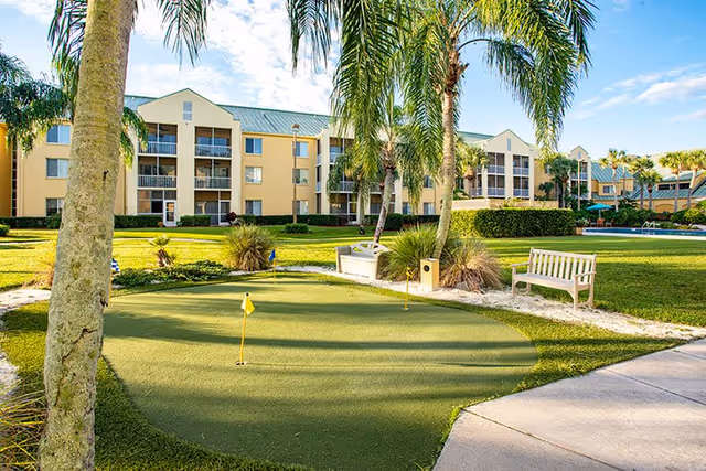 Outdoor view of The Fountains of Melbourne senior living facility featuring a small putting green with flags, palm trees, benches, and a well-maintained lawn with the building in the background under a partly cloudy sky.