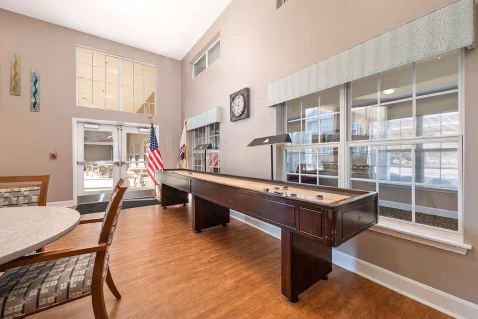 A bright indoor recreational room with a shuffleboard table, round table with chairs, large windows, and double glass doors leading outside. The room has light-colored walls, wood flooring, and an American flag near the doors.