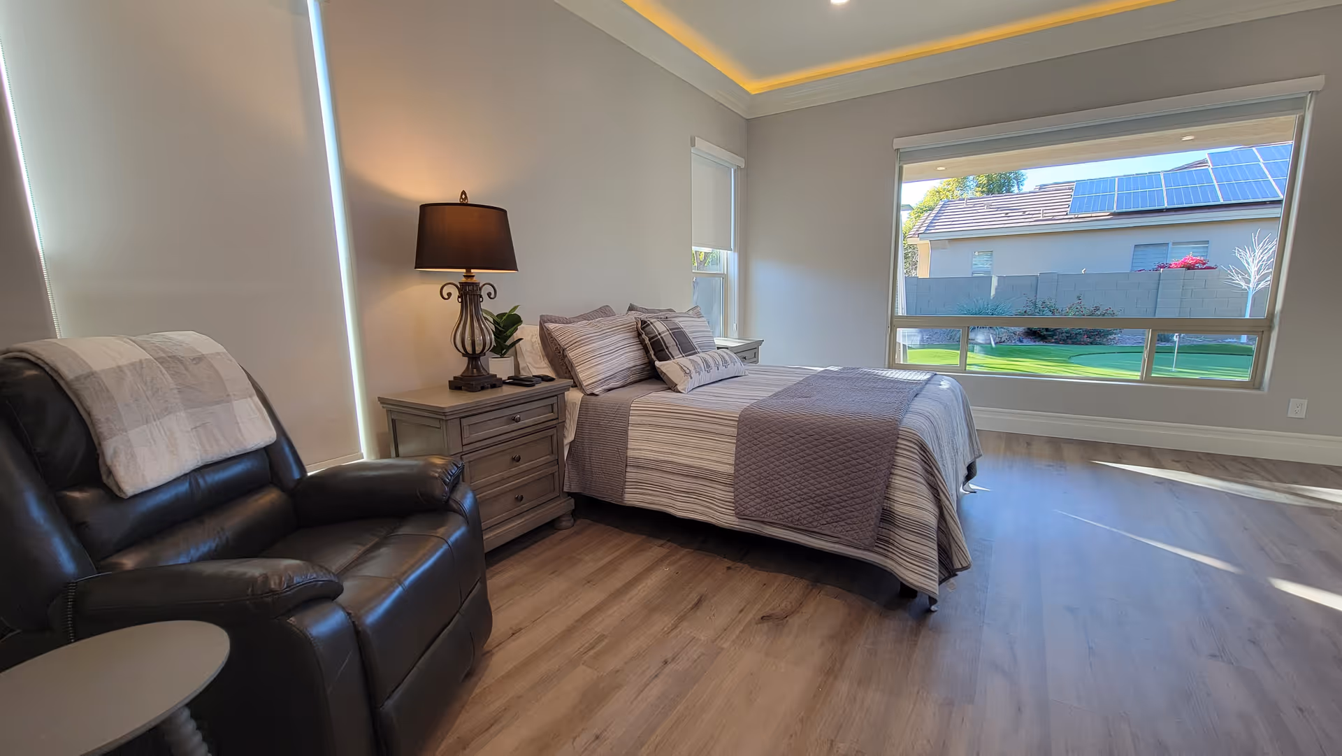 A bright and spacious bedroom with a large window showing a green lawn outside. The room features a neatly made bed with striped bedding and a gray quilt, a wooden nightstand with a decorative lamp and plant, and a black leather recliner chair with a plaid blanket draped over the back. The floor is wooden, and the walls are painted light gray.