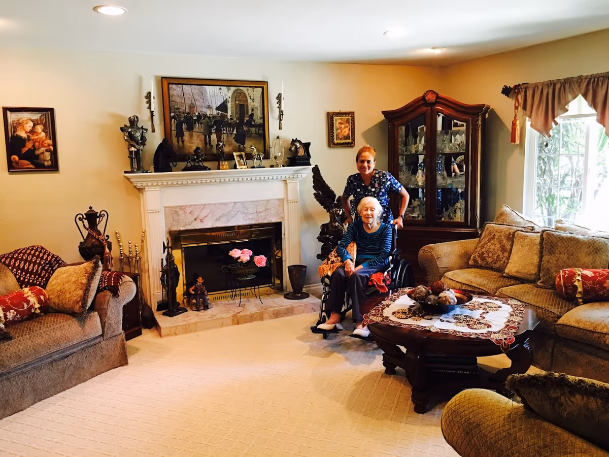 A cozy living room with beige walls and carpet, featuring a fireplace decorated with various sculptures and framed paintings above it. There are two large sofas with patterned cushions, a wooden coffee table with a decorative cloth and centerpiece, and a glass cabinet filled with glassware. An elderly woman in a wheelchair is seated near the fireplace, accompanied by a caregiver standing behind her. Natural light comes in through a window with curtains on the right side.