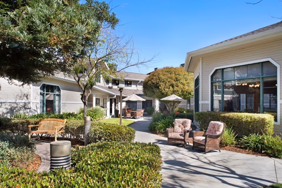 Outdoor courtyard area at Ivy Park at Salinas featuring a paved walkway, green shrubs, trees, a wooden bench, two cushioned wicker chairs, and patio tables with umbrellas. The buildings surrounding the courtyard have large windows and beige siding under a clear blue sky.